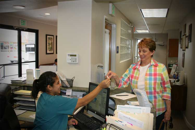 A dental clinic staff in a blue scrubs talking to a patient in colorful plaid top in a reception area