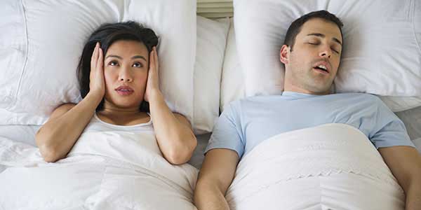 A female covering her ears with her hands  and beside her is a male sleeping with an open mouth, snoring
