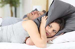A female covering her ears with a grey pillow and beside her is a male sleeping with an open mouth, snoring
